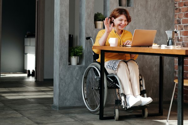 Photo of a woman sitting in a wheelchair smiling waving and using a laptop