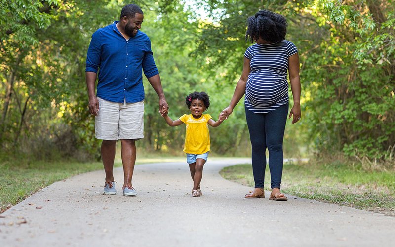 A family of three walk on a path in a woodland area. Mum and Dad are both smiling down at a little girl between them all smiling.