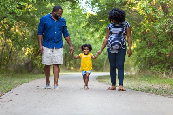Image shows a family walking among trees all holding hands. There's dad, a toddler in the middle and mum on the right. Mum is pregnant. They are all looking at the toddler and smiling.