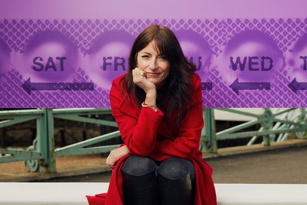 Davina McCall in a red coat sitting and facing camera with head in hand. Behind her is a big billboard of a purple pill packet.