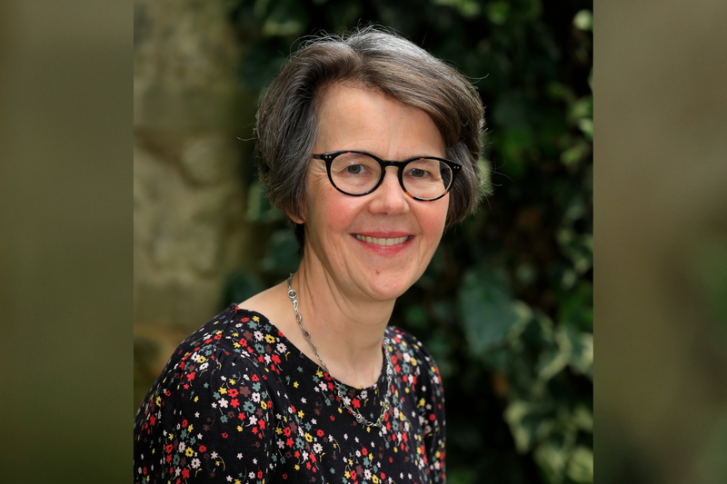 A white woman with short grey hair and glasses. She's smiling and wearing a black top with colourful floral print.
