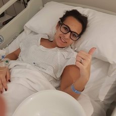 A young white women lying in a hospital bed, she is giving thumbs up