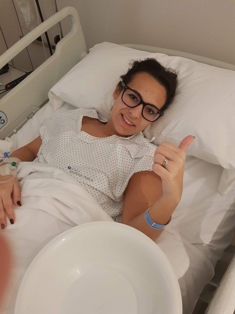 A young white women lying in a hospital bed, she is giving thumbs up