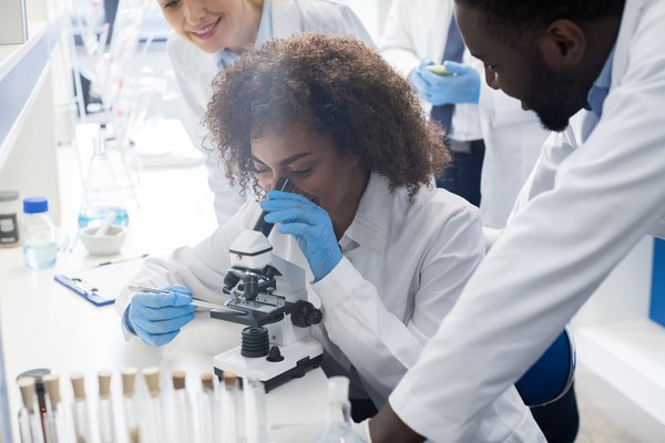 A Black female researcher is looking into a microscope in a laboratory. Two other researchers can be seen looking on. The body of a third researcher is in the background
