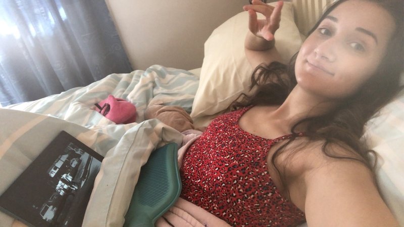 A young white women, lying in bed with a blue hot water bottle on her stomach