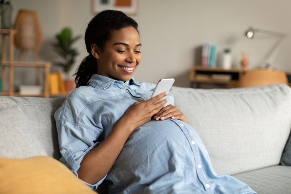 A pregnant Black women in a loose fitting blue shirt is sitting on a sofa with her left hand on her bump and her phone in her right hand. She is looking at her phone and smiling