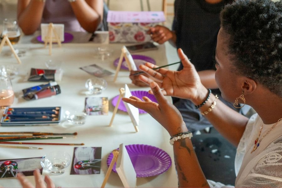A table showing art supplies with a side view of a Black woman holding her paintbrush gesturing in an animated way