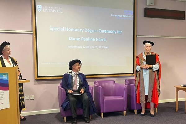 Dame Pauline Harris receiving her specialist honorary degree. She is standing wearing her graduation gown and cap on the left of her is a man sat on a chair and lady stood at a lectern. Behind them is a screen announcing the degree.