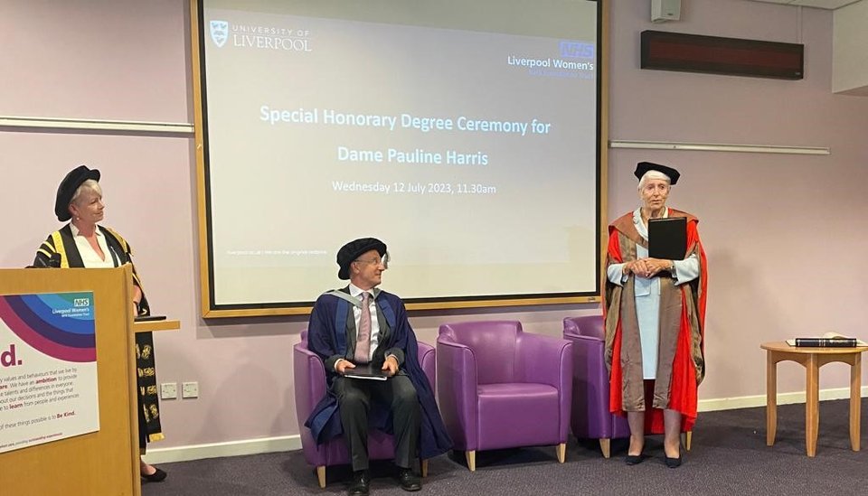 Dame Pauline Harris receiving her specialist honorary degree. She is standing wearing her graduation gown and cap on the left of her is a man sat on a chair and lady stood at a lectern. Behind them is a screen announcing the degree.