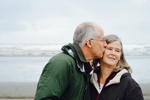 Image shows older couple on a beach. Man is kissing woman's cheek.