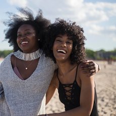Image shows two women with their arms around each other, smiling. They are on a beach.