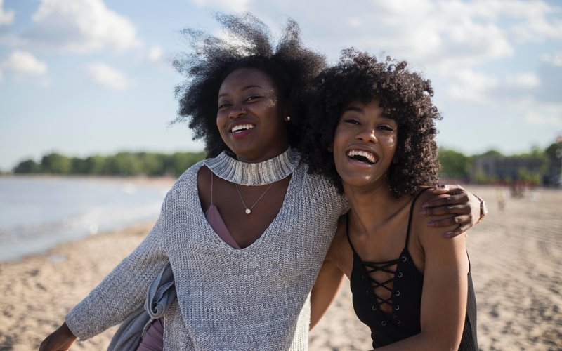 Image shows two women with their arms around each other, smiling. They are on a beach.
